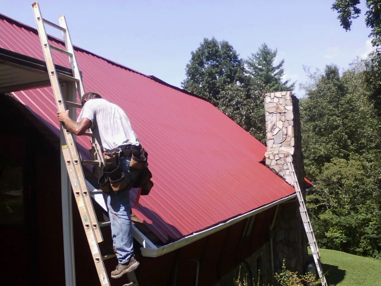 Roofer on a ladder beside a red metal roof and stone chimney — Soffit & Fascia Repair by Roofing Brothers in Croton On Hudson